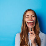 Brunette woman smiles as she holds a magnifying glass up to her teeth after professional teeth whitening