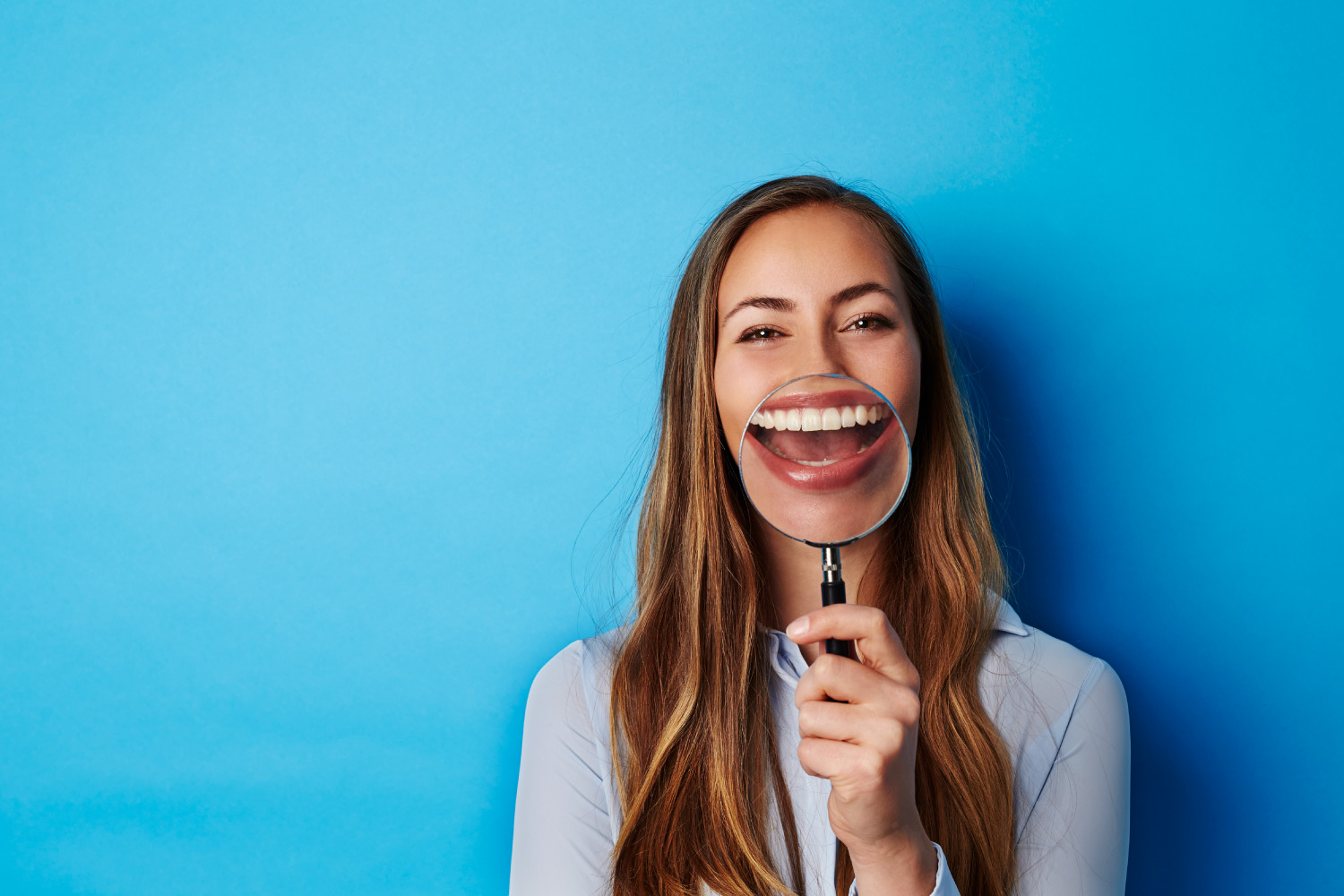 Brunette woman smiles as she holds a magnifying glass up to her teeth after professional teeth whitening
