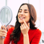 Brunette woman in a red blouse smiles and points to her teeth as she looks in a handheld mirror