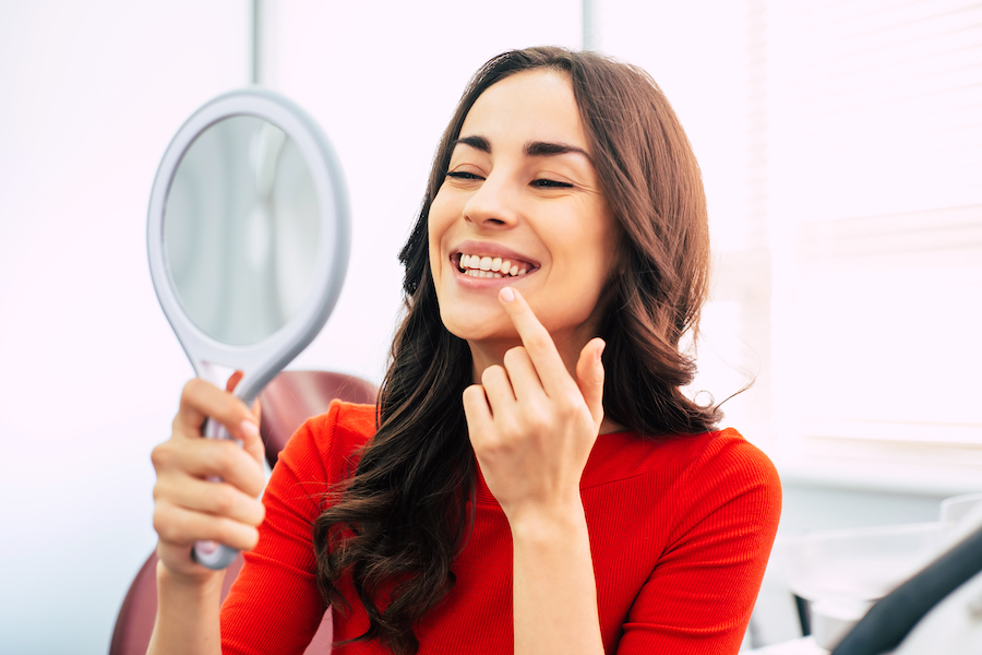 Brunette woman in a red blouse smiles and points to her teeth as she looks in a handheld mirror