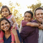 Mom and dad give piggyback rides to their son and daughter as they smile outside