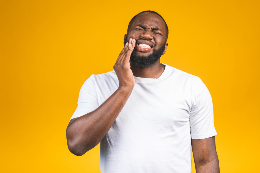 Black man in a white t-shirt cringes in pain and touches his cheek because of an emergency toothache